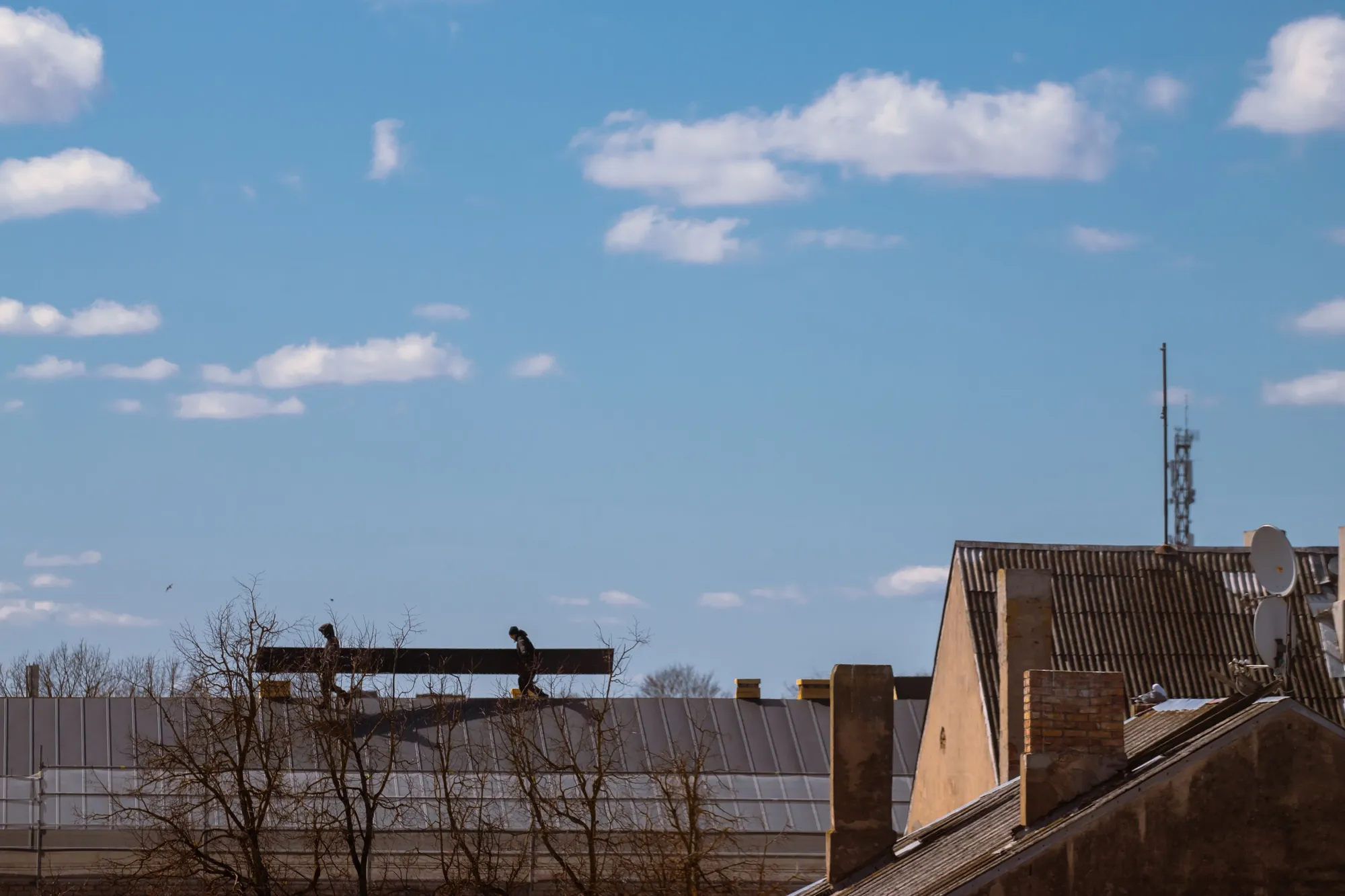A roofscape under a blue sky, two workers carrying a beam along a metal roof
